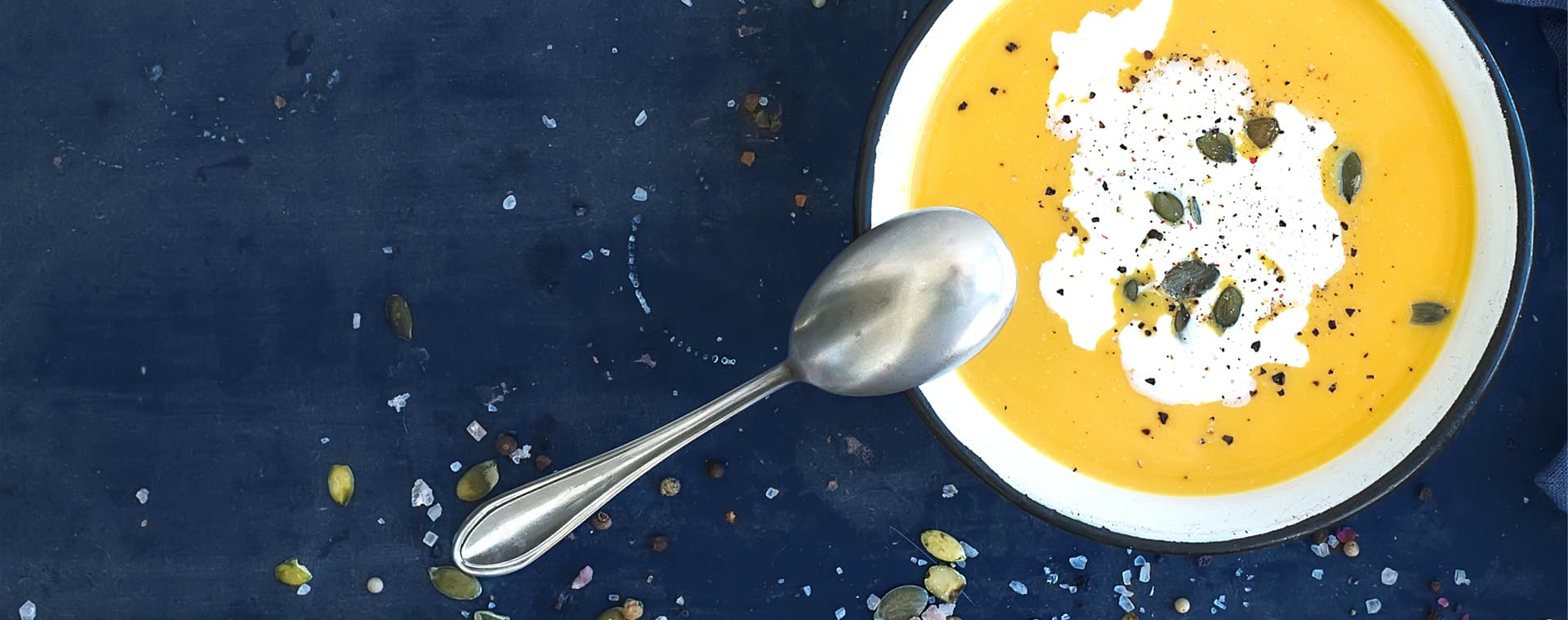 A photo of a bowl of cold pumpkin soup with sour cream, sprinkled with pepitas and freshly ground black pepper.
