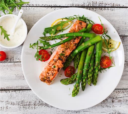 A photo of a dish of grilled salmon and asparagus, halved fresh cherry tomatoes, with mixed greens and lemon wedges.