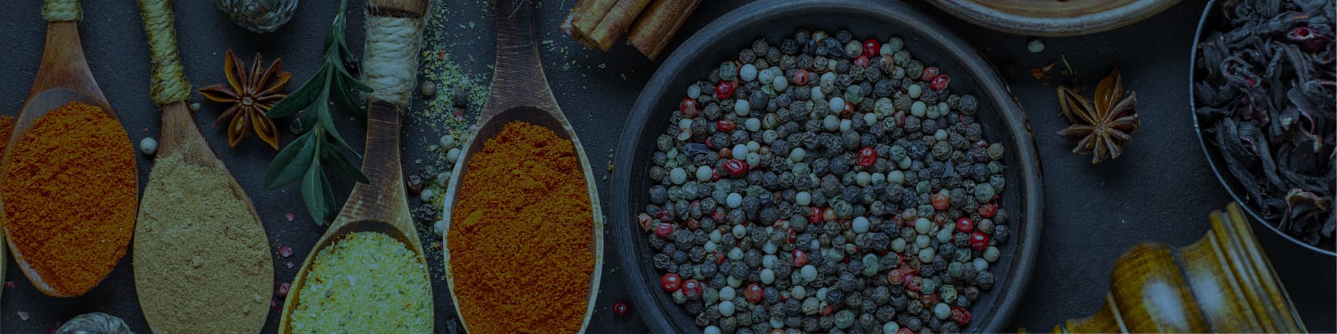 A photo of an assortment of freshly ground turmeric, cardamom, ginger and cumin, assortment of red, black, white and green peppercorns in a bowl, dried ancho chiles in a bowl, surrounded with pices of star of anise and cinnamon sticks ontop of a kitchen countertop.