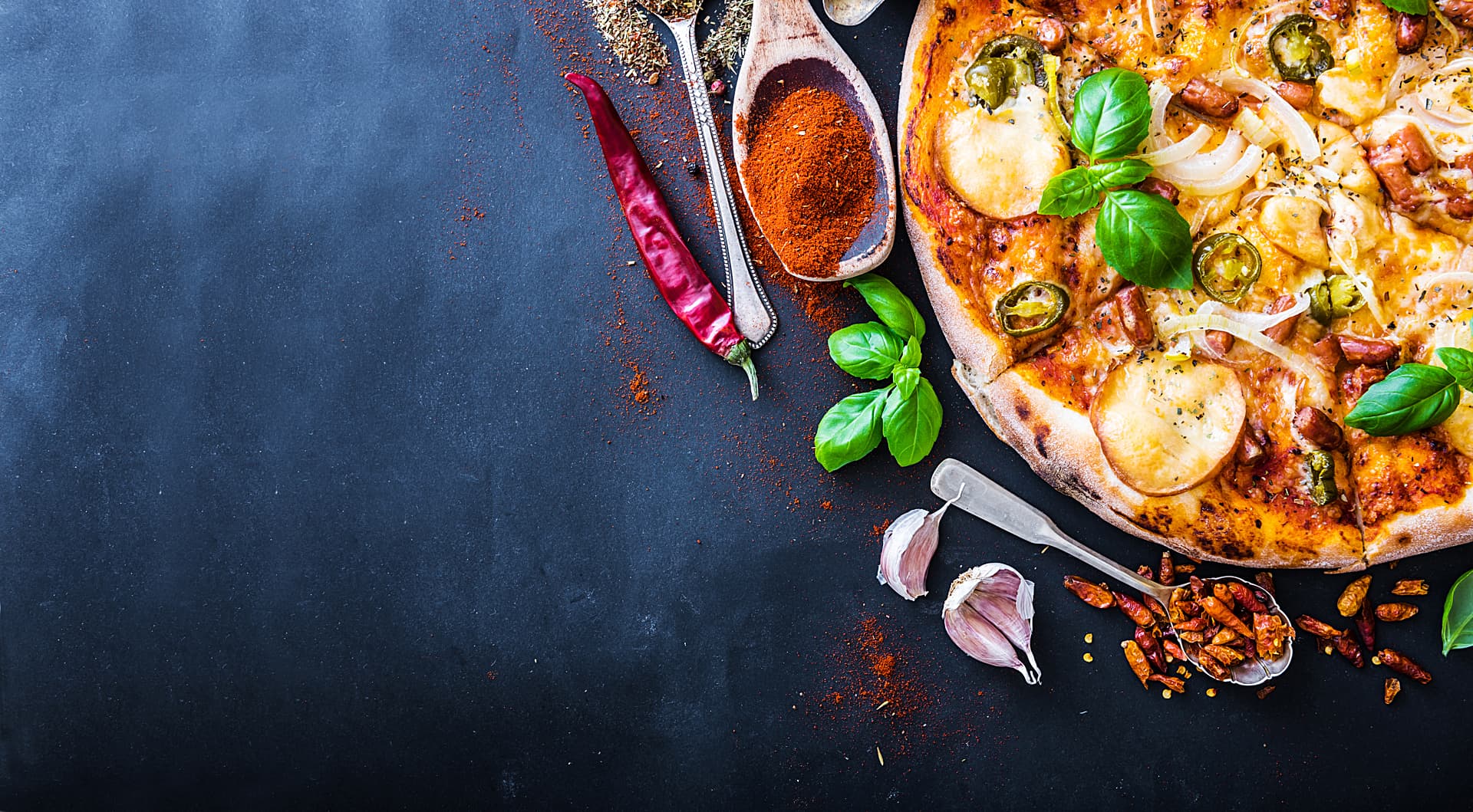 A photo of a freshly baked sausage, potato and green Italian long hot peppers topped with fresh basil, and surrounded with an assortment of fresh and dried Calabrian chili peppers, dried red chili peppers, cloves of garlic, ground cayenne and fresh basil leaves.
